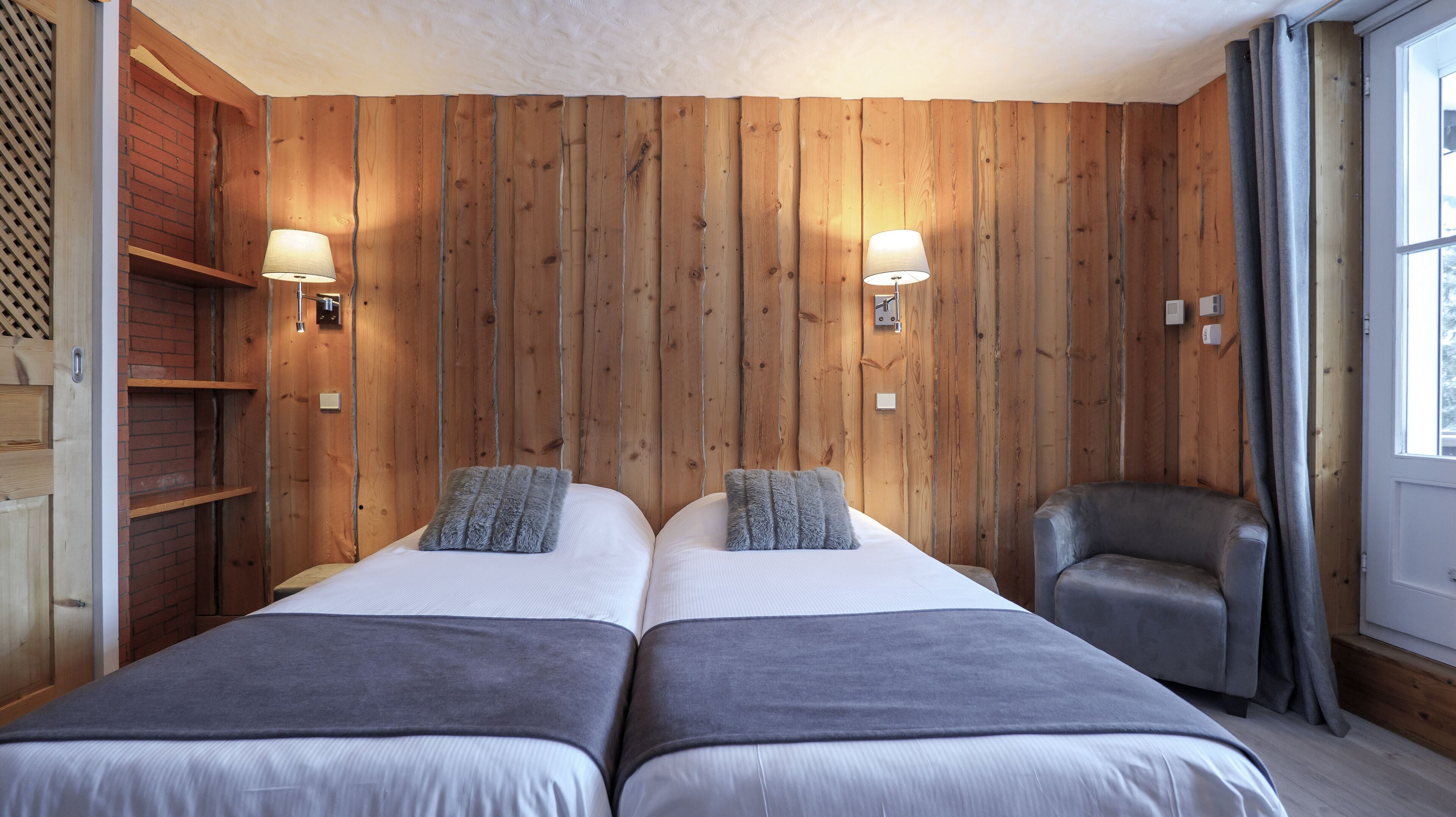 Una habitación con dos camas, paredes de madera, una silla gris y dos lámparas fijadas a la pared.