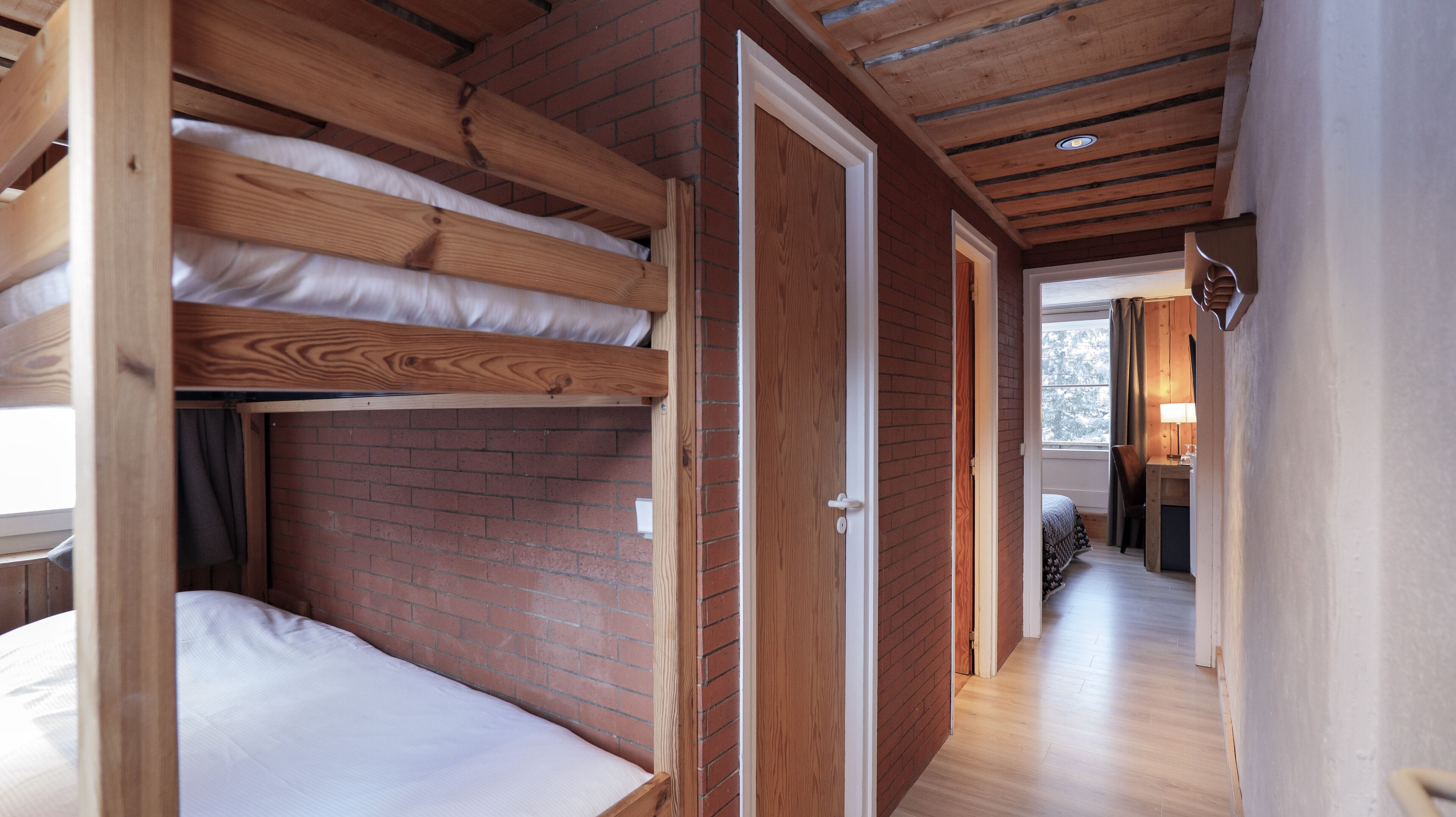 A room with bunk beds, a brick wall, wooden beams, a door, and a hallway leading to a kitchen area.