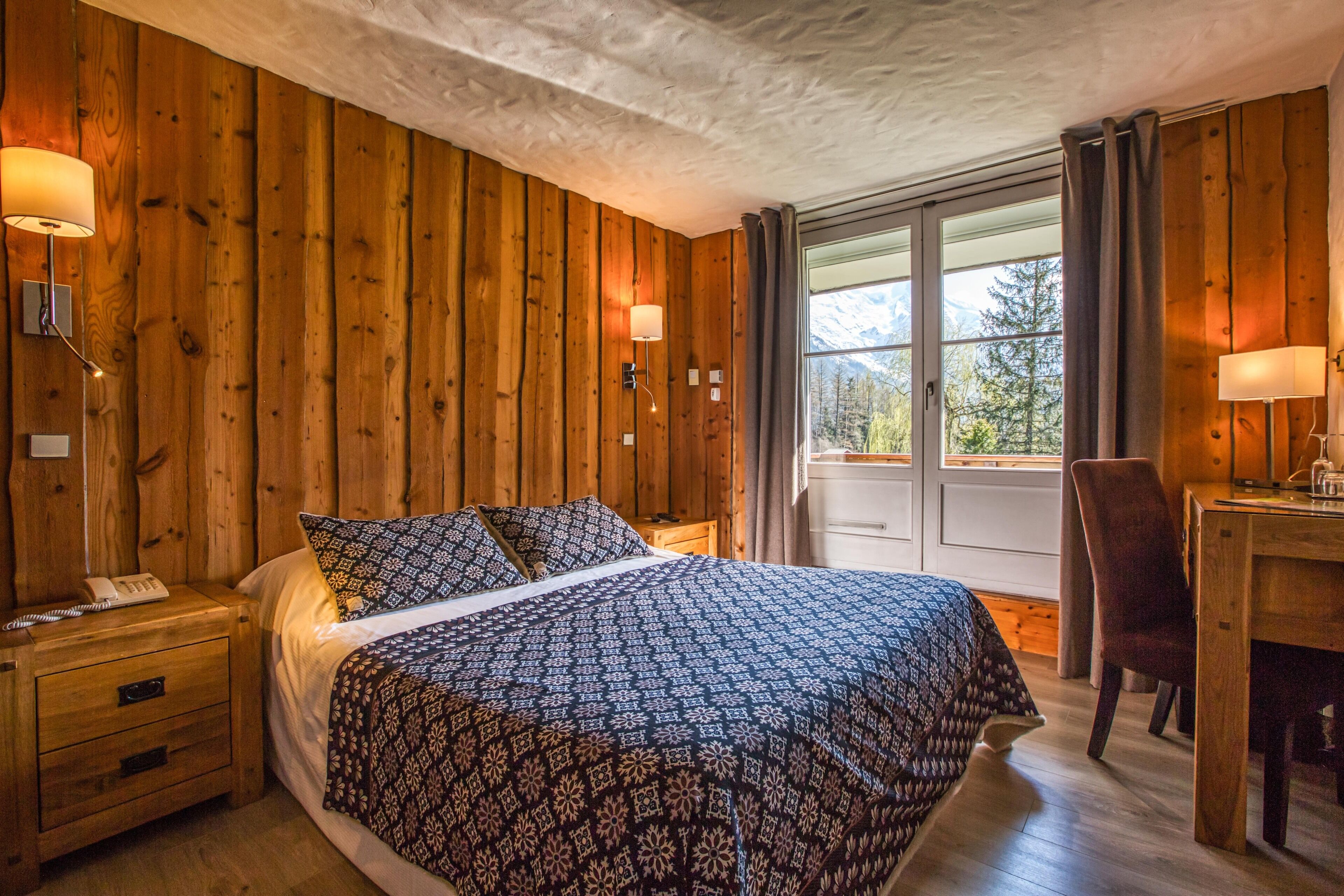 A wooden-paneled bedroom with a large bed, bedside lamps, a desk, and a window with a mountain view.