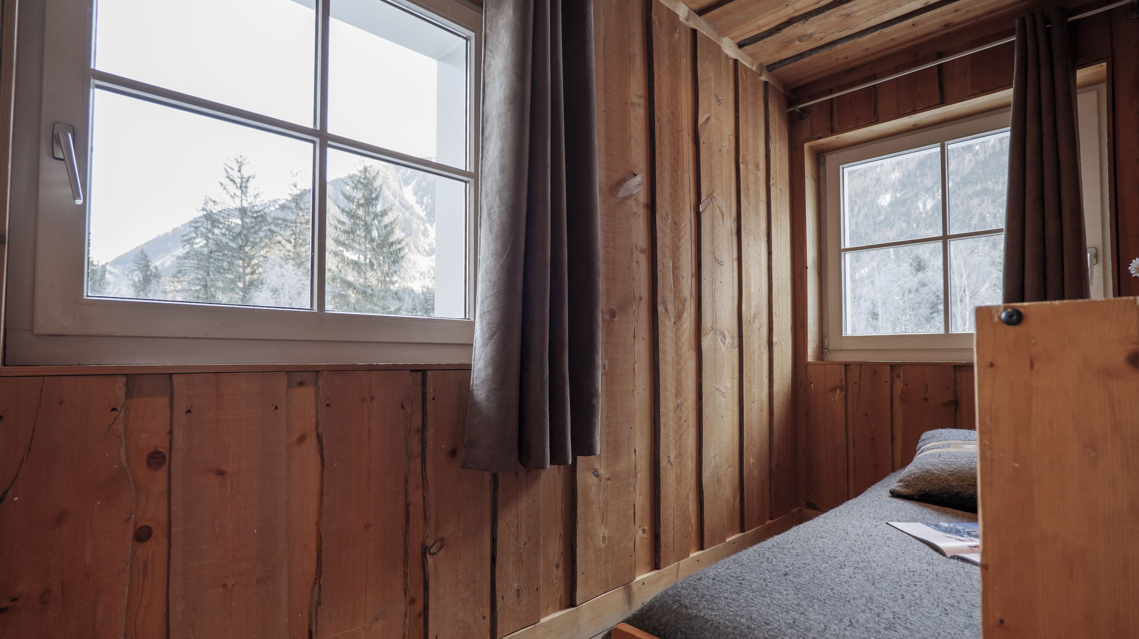 A cabin with wooden walls, two windows with curtains, a bed, and a view of snow-covered mountains.
