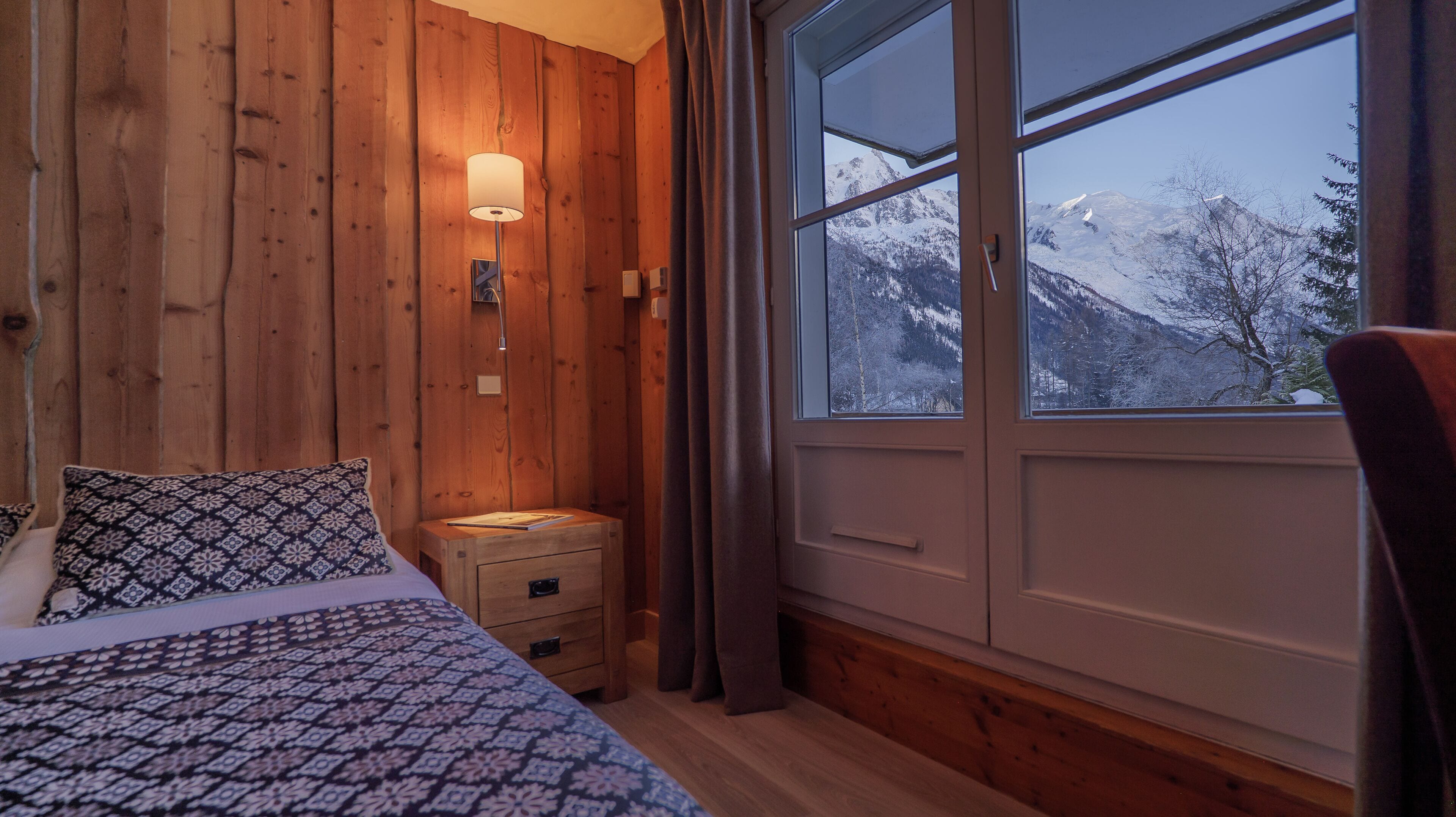 A wooden room with a bed, a bedside table, a lamp, and a window with a snowy mountain view.