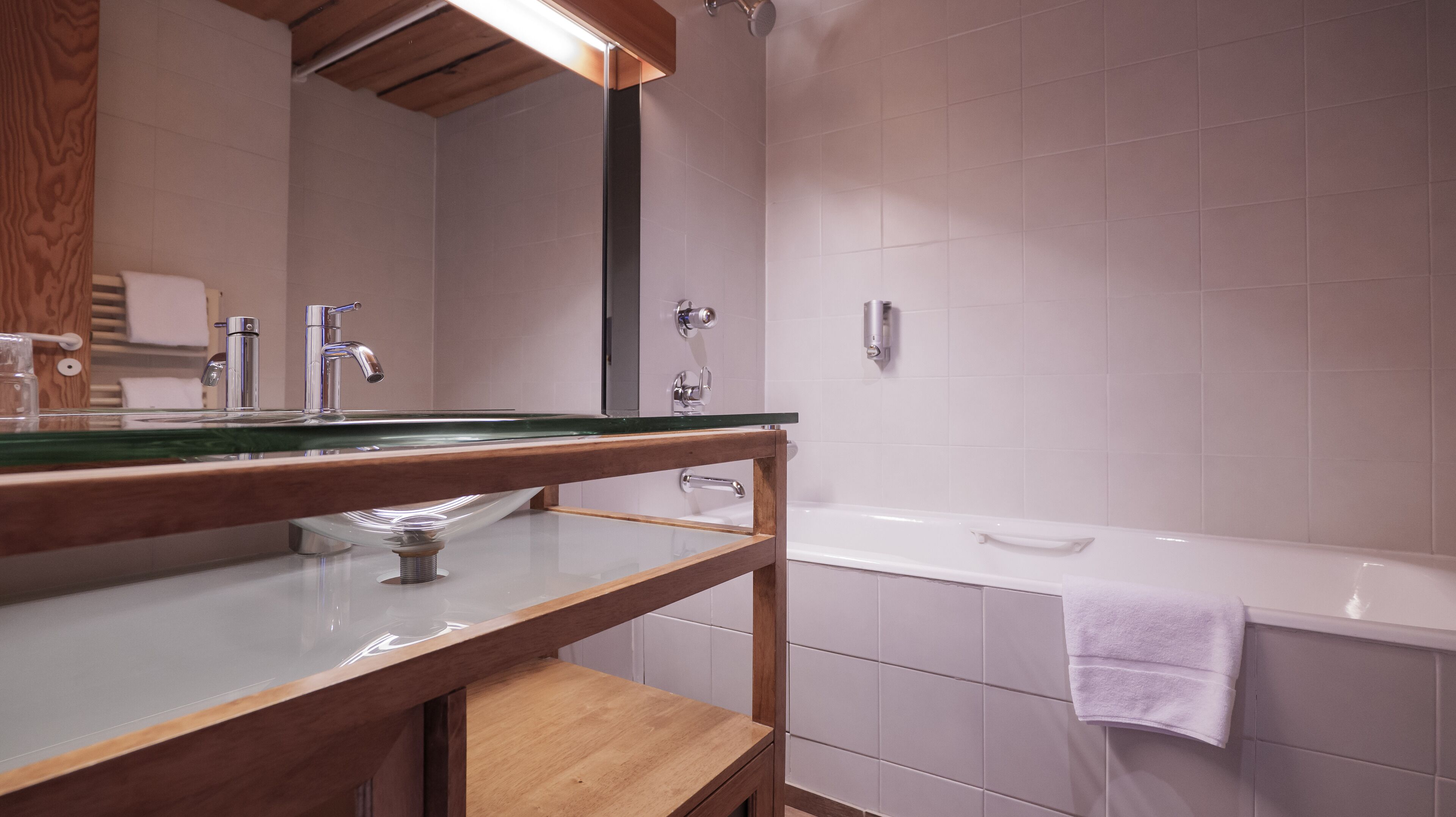 A bathroom with a wooden vanity, a glass sink, a mirror, a bathtub, and a towel hanging on the wall.