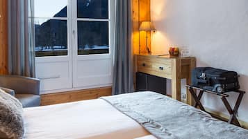 Habitación de hotel con cama, escritorio, silla, maleta sobre carrito y ventana con vistas a árboles y montañas.