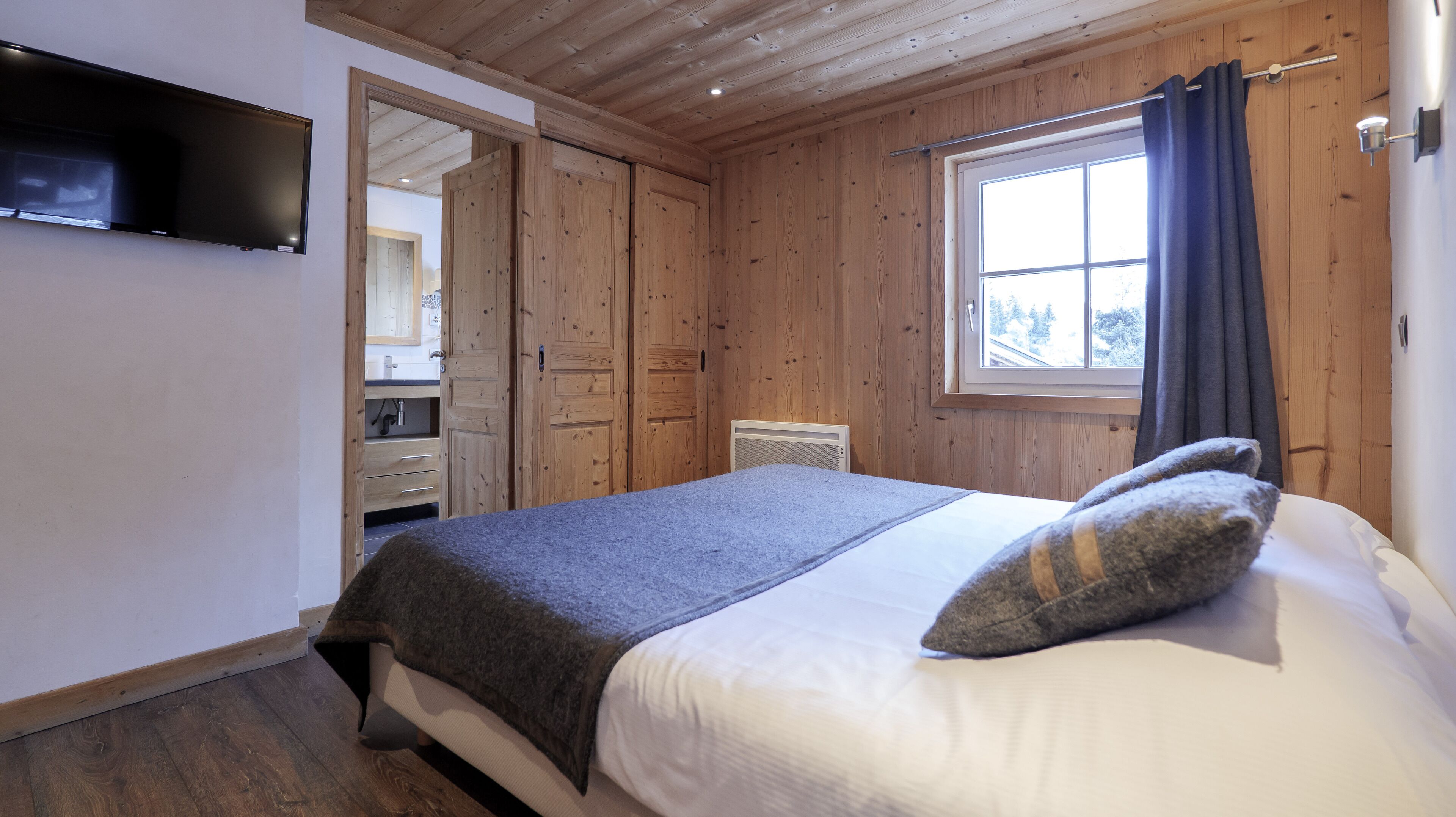 Un dormitorio con cama, televisión montada en la pared, paneles de madera, ventana con cortinas y baño visible a través de un umbral abierto.