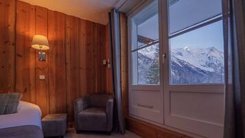 Una habitación con paredes de madera, un ventanal con vista a la montaña, una cama, un sillón y una lámpara.