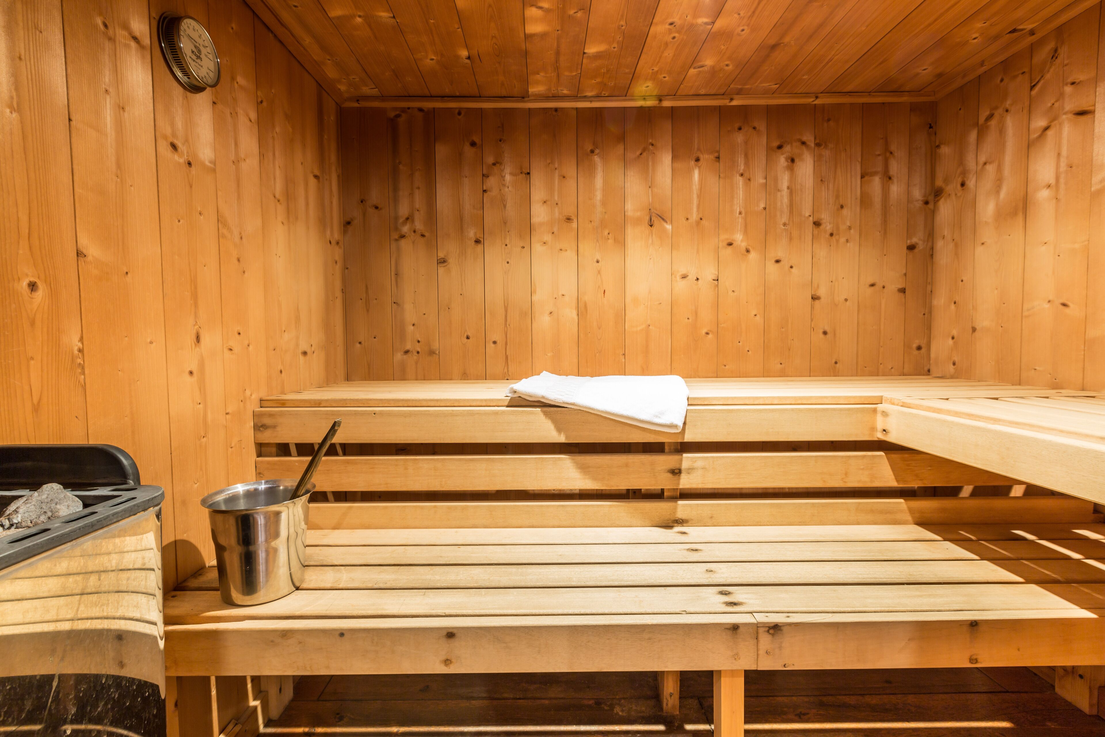 Una sala de sauna con bancos de madera, un cubo metálico con una cuchara y un reloj en la pared.