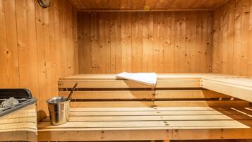 Una sala de sauna con bancos de madera, un cubo metálico con una cuchara y un reloj en la pared.