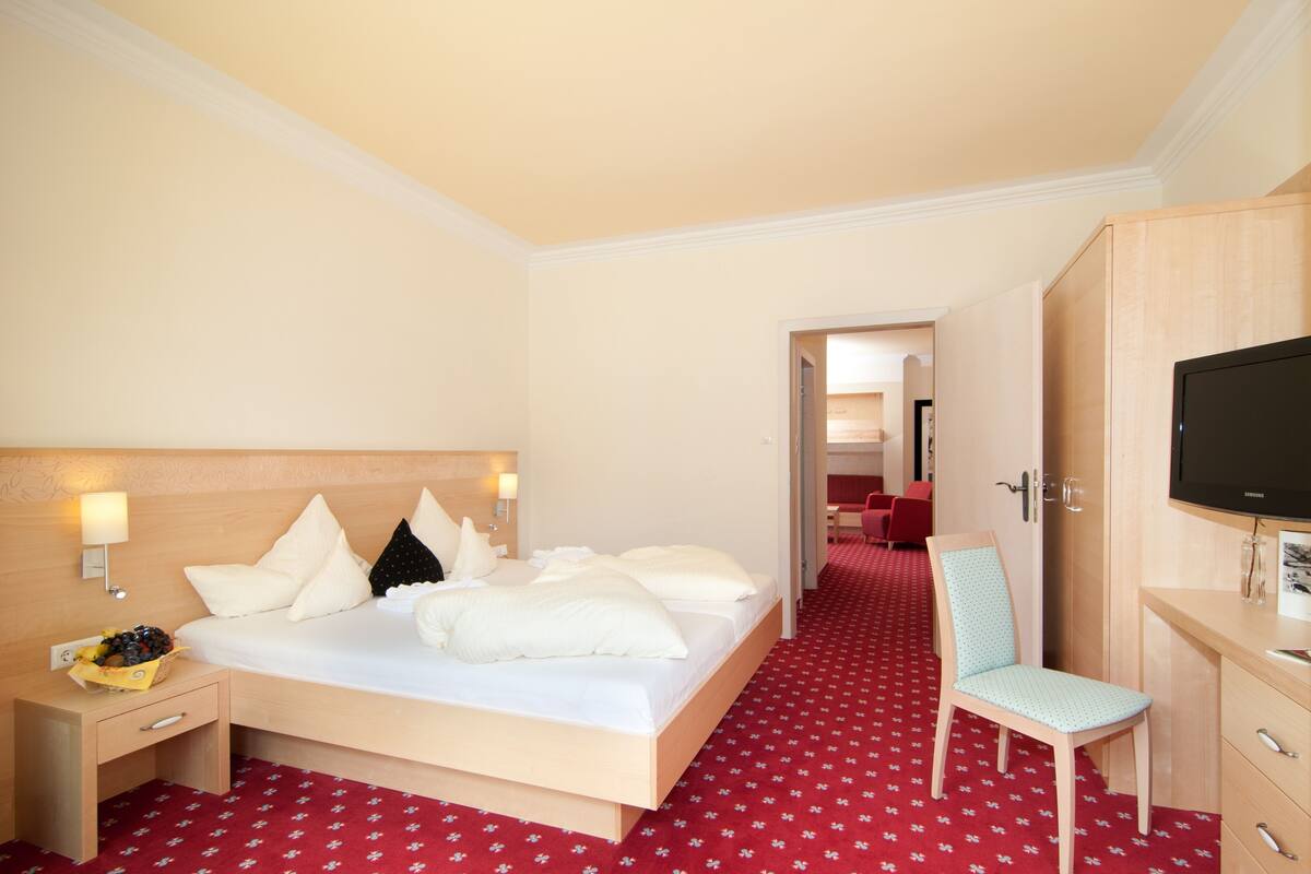 Une chambre d’hôtel comprenant un lit, une table de chevet, une chaise, un bureau, une télévision et un sol recouvert d’une moquette rouge.