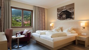 Hotelkamer met een groot bed, een tafeltje met een stoel, uitzicht op een berglandschap en een schilderij aan de muur.