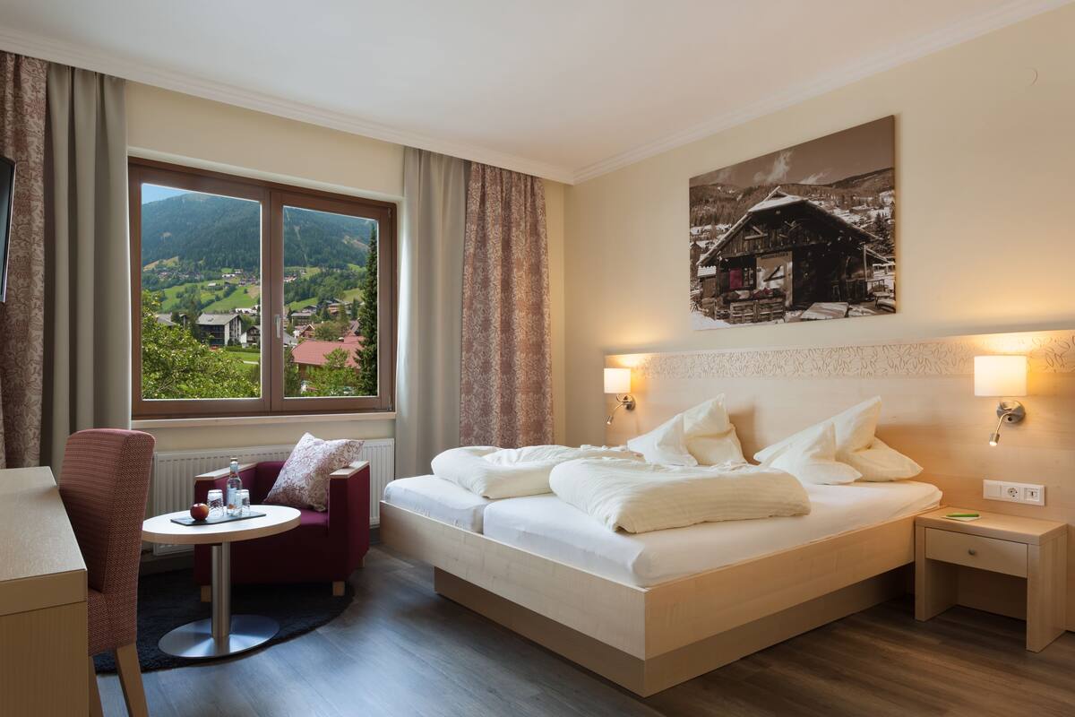 Une chambre d’hôtel avec un grand lit, une petite table avec une chaise, une vue sur un paysage montagneux et un tableau au mur.