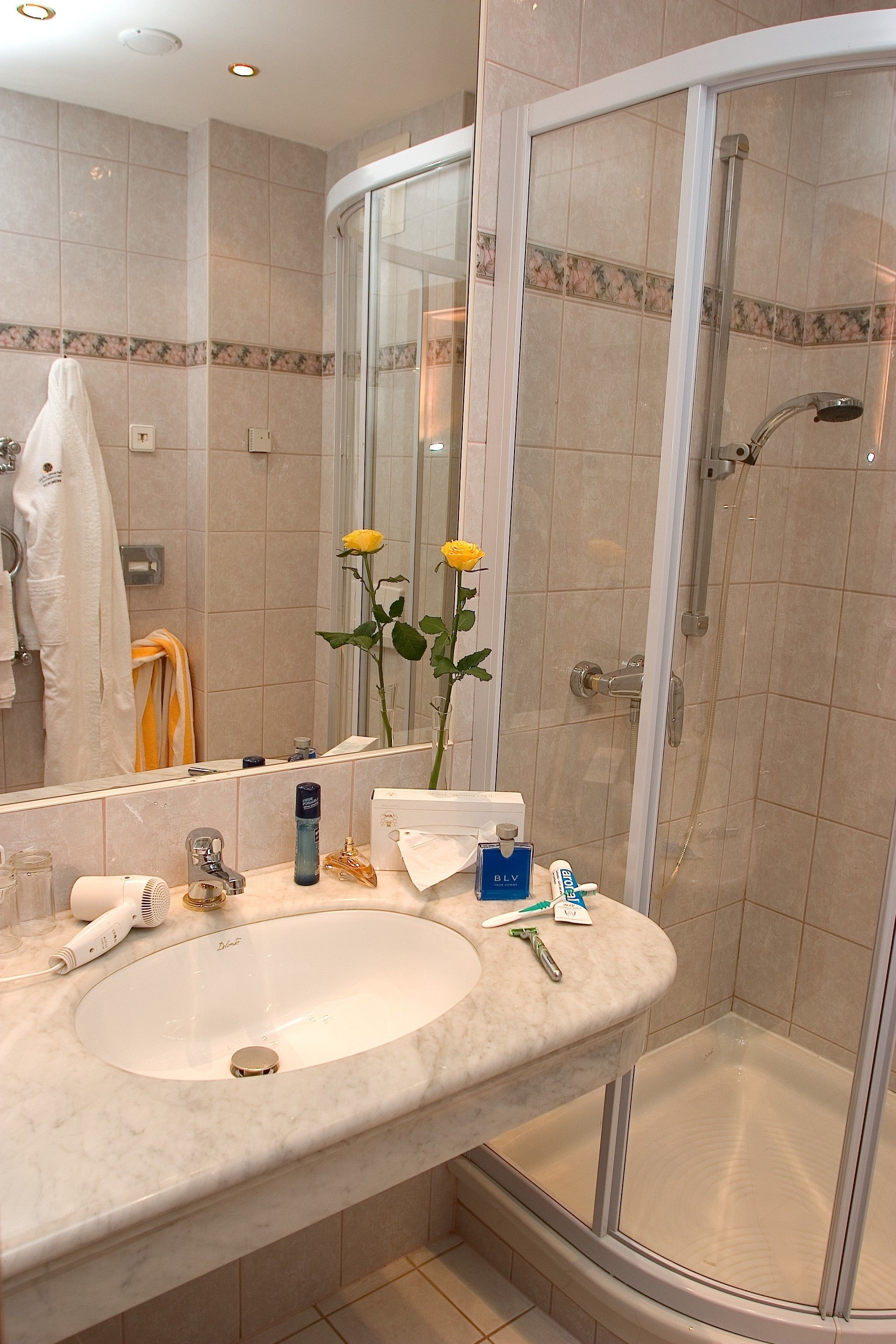 A bathroom with a white sink, a glass shower enclosure, and a mirror reflecting a yellow rose.