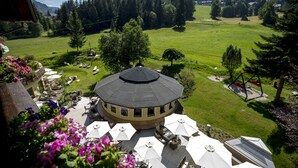 Een rond gebouw met dak, omringd door terraszitplaatsen met parasols, gelegen voor een groene weide en bomen.
