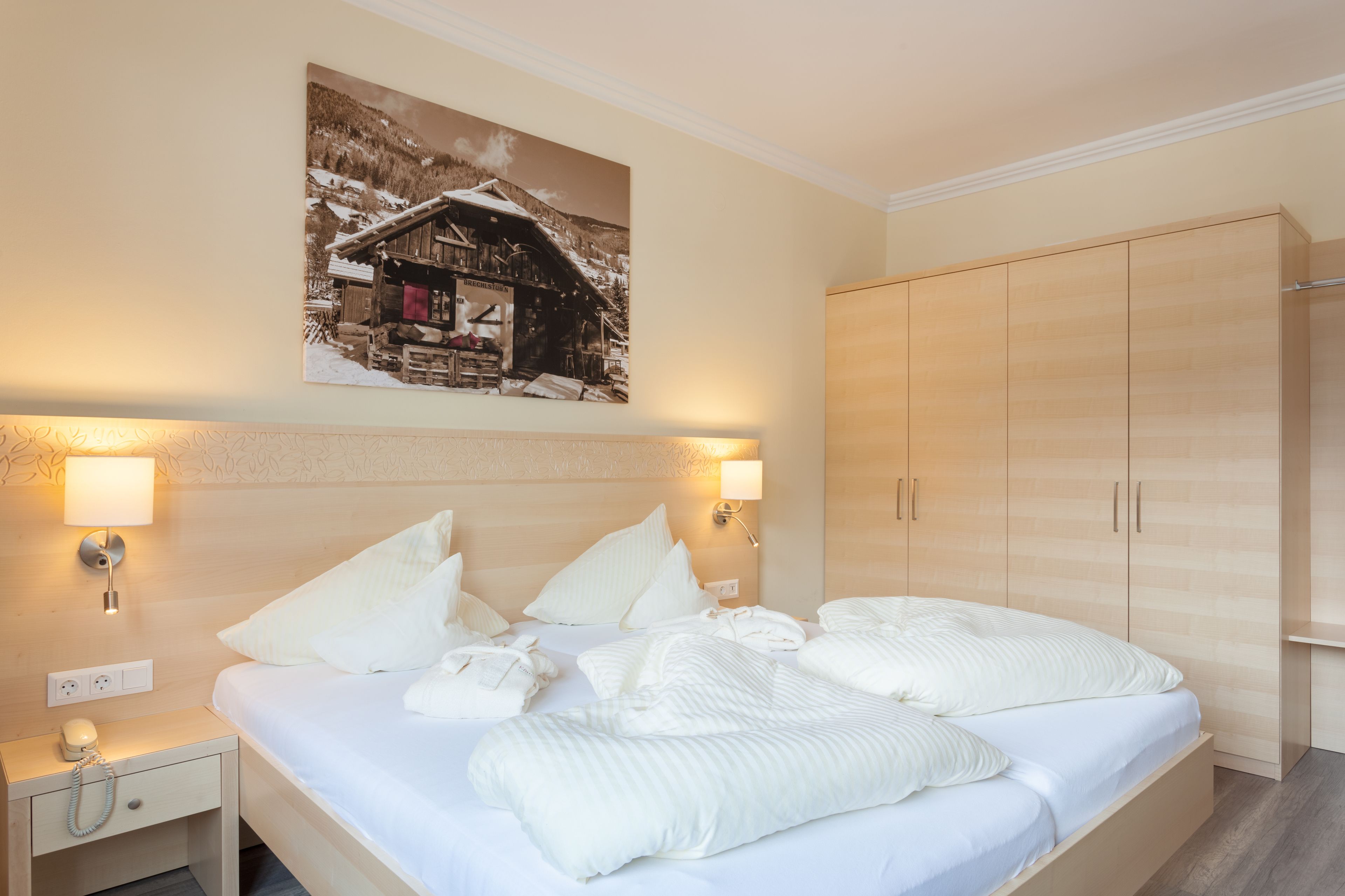 Habitación de hotel con cama, lámparas de noche, mesita de noche, armario de madera y un cuadro colgado en la pared que muestra un paisaje nevado de montaña.