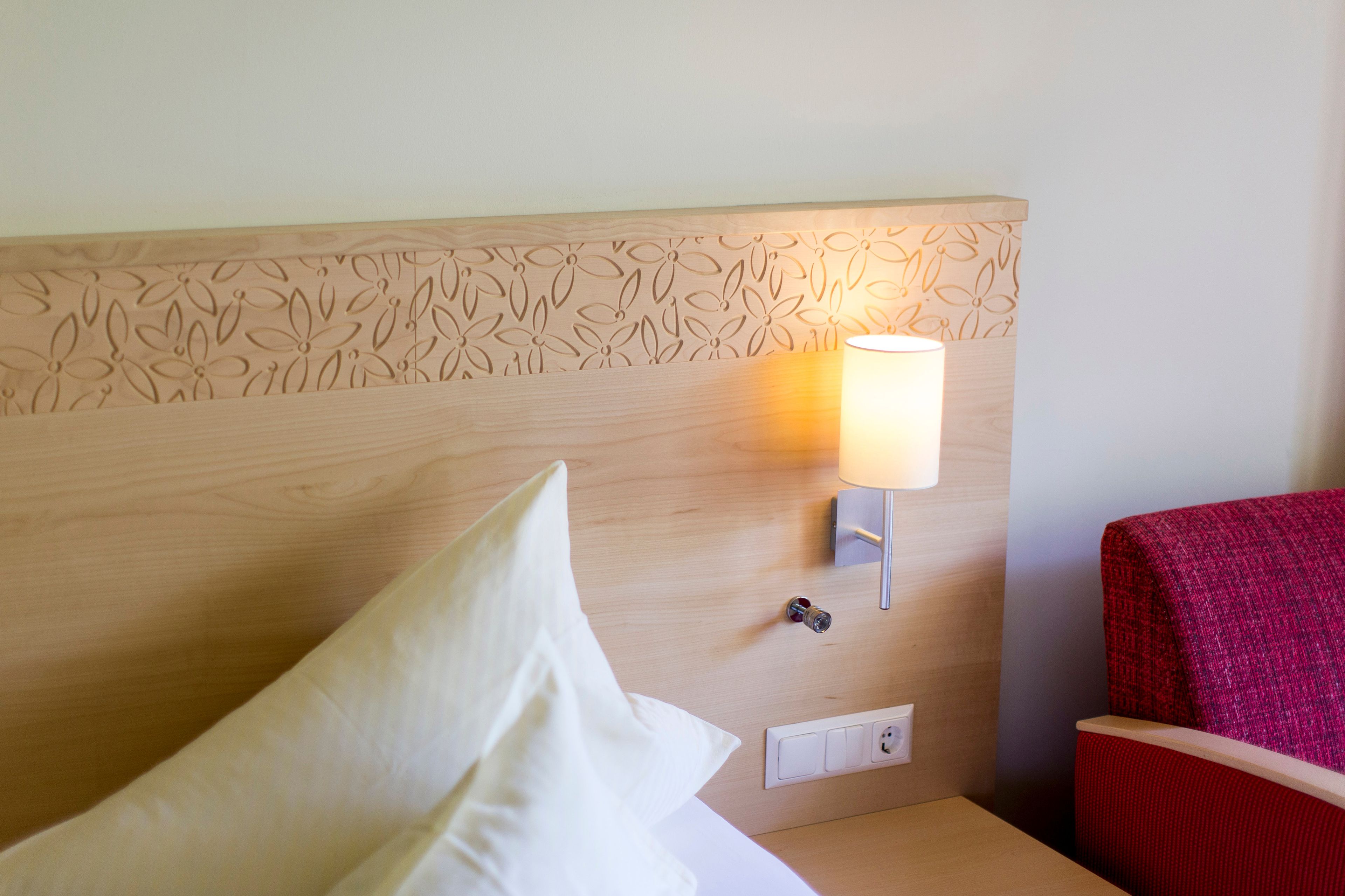 A hotel room with a wooden headboard, a bedside lamp, a red chair, and a white pillow.