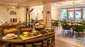 Een ontbijtbuffet met diverse broden en gebak, een zithoek met groene stoelen, en een groot raam met uitzicht op het omliggende landschap.