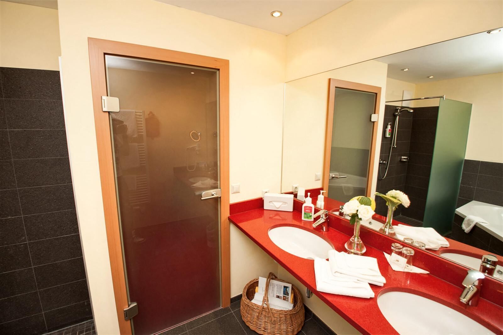 Un baño moderno con encimera roja, dos lavamanos, un espejo grande y zona de ducha.