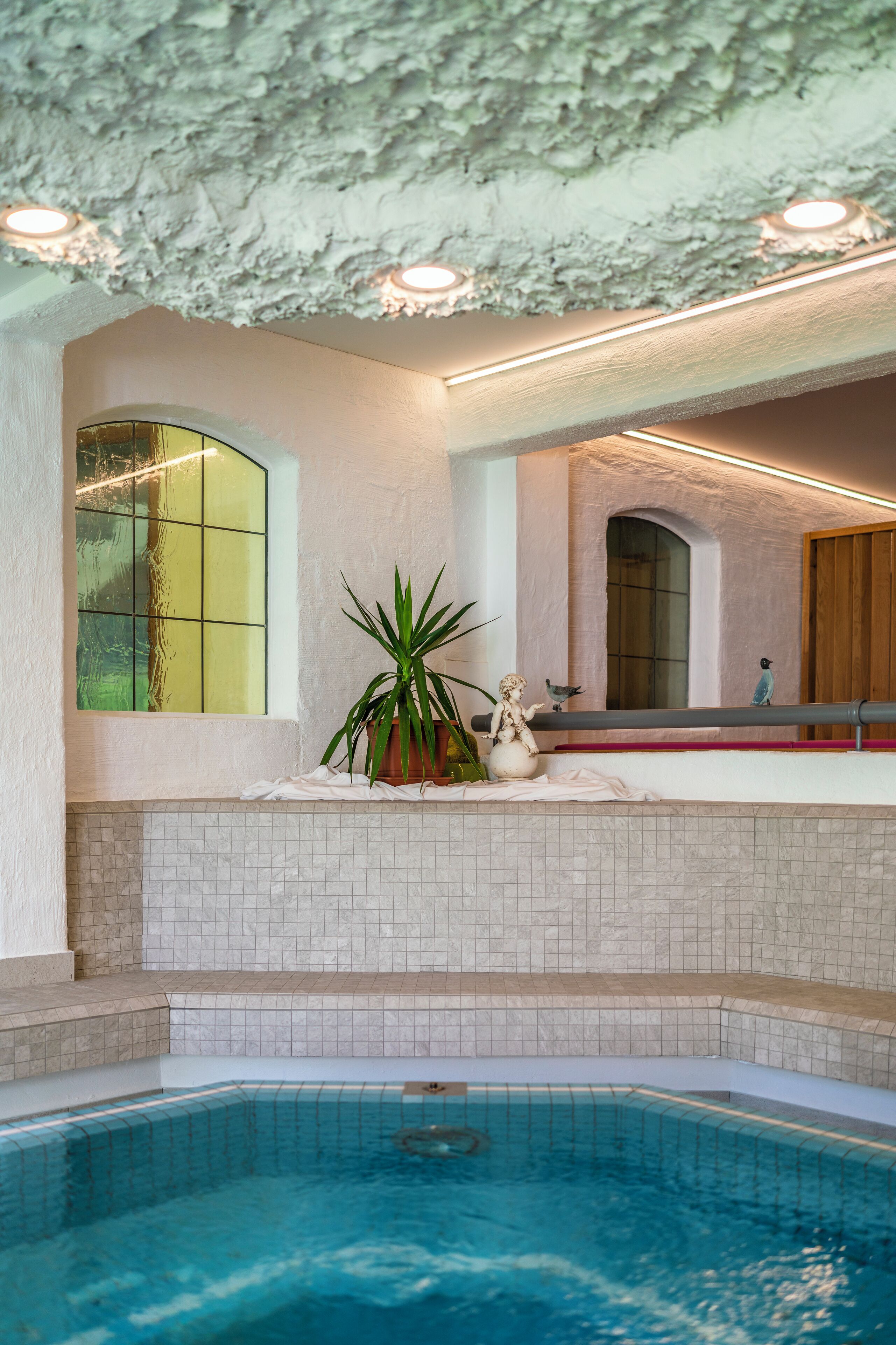 An indoor swimming pool with tiled walls, a ceiling with textured plaster, and a window with a green tint.