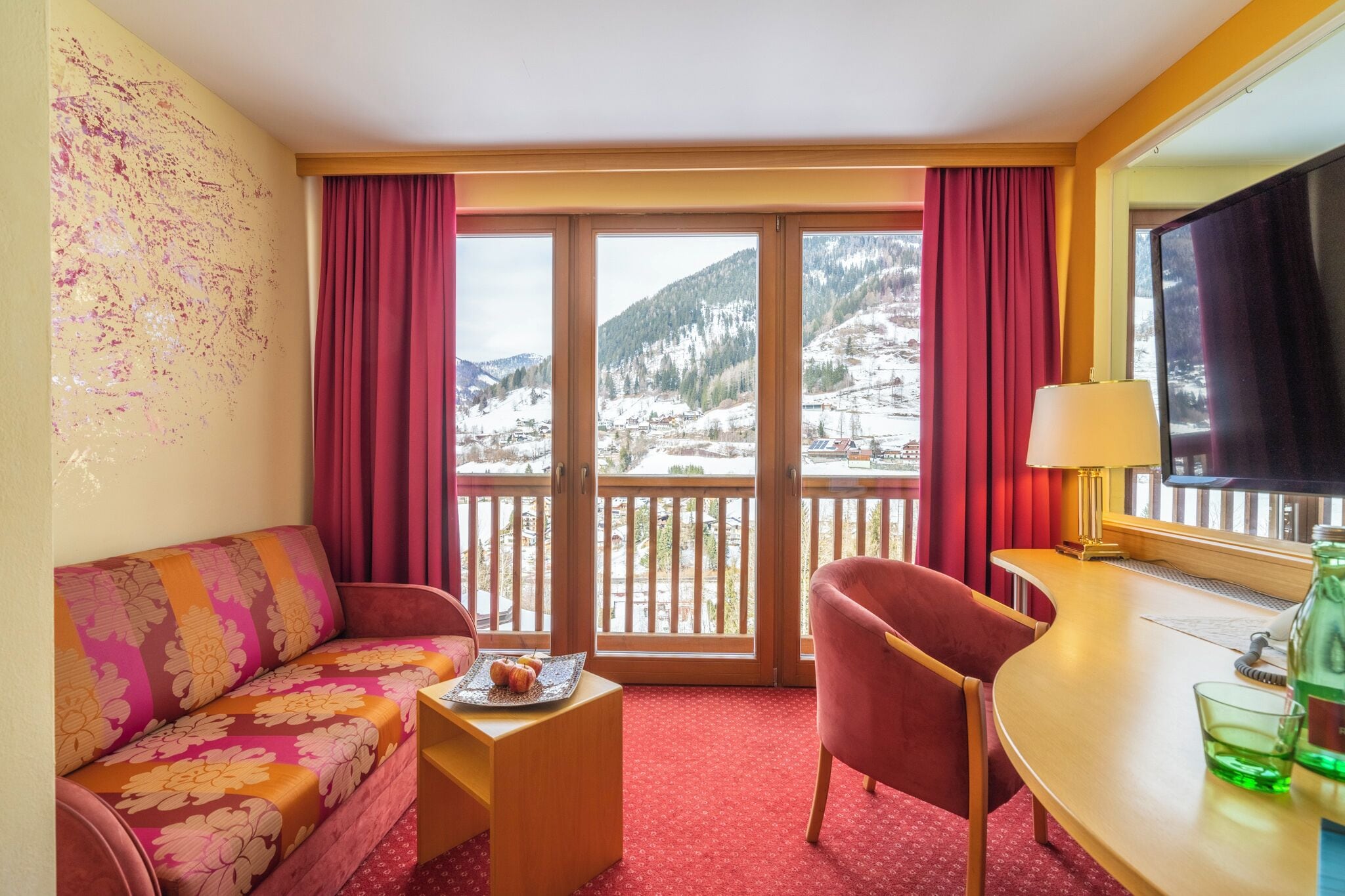 A hotel room with a sofa, chair, small table, and a balcony with a view of snow-covered mountains.