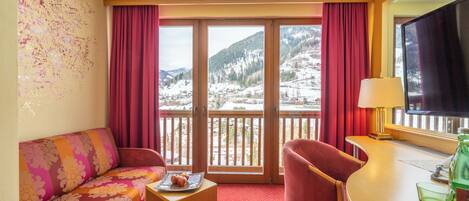 A hotel room with a sofa, chair, small table, and a balcony with a view of snow-covered mountains.