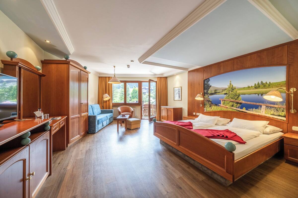 Una habitación amplia con una cama grande, zona de estar con sofá y sillones, televisor, armarios de madera y un mural decorativo en la pared.
