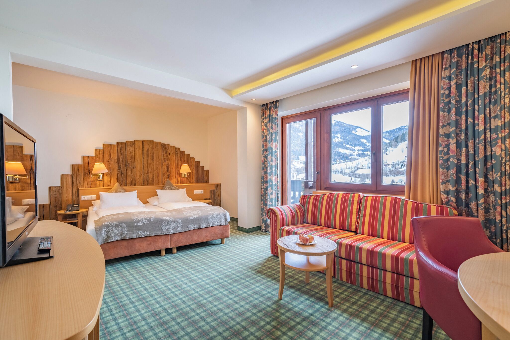 Habitación de hotel con cama, sofá, mesa y un amplio ventanal con vistas a montañas.