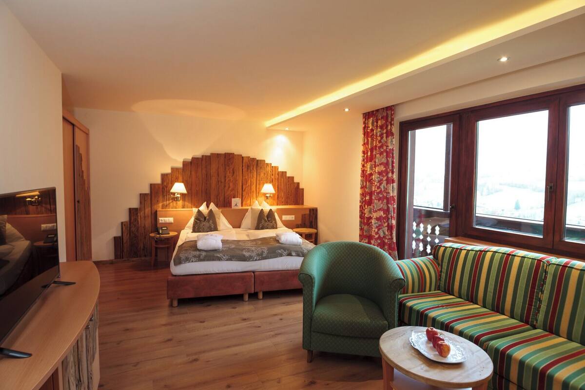 Un cuarto de hotel con una cama, un sillón verde, un sofá a rayas, un escritorio de madera y un ventanal con vistas.