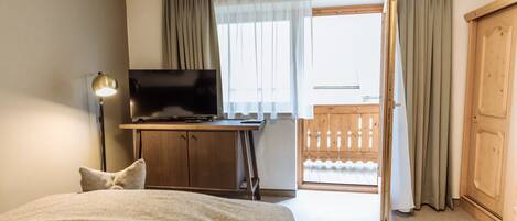 A hotel room with a bed, a desk with a TV, a wooden wardrobe, and a balcony with curtains.