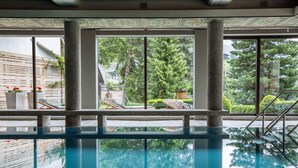 An indoor swimming pool with clear glass walls, stone columns, and a view of a landscaped garden. - Nationalpark Lodge Grossglockner (Heiligenblut)