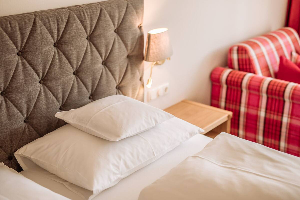 A neatly made bed with white linens and a tufted headboard, a bedside lamp, and a plaid blanket on a red and white chair.