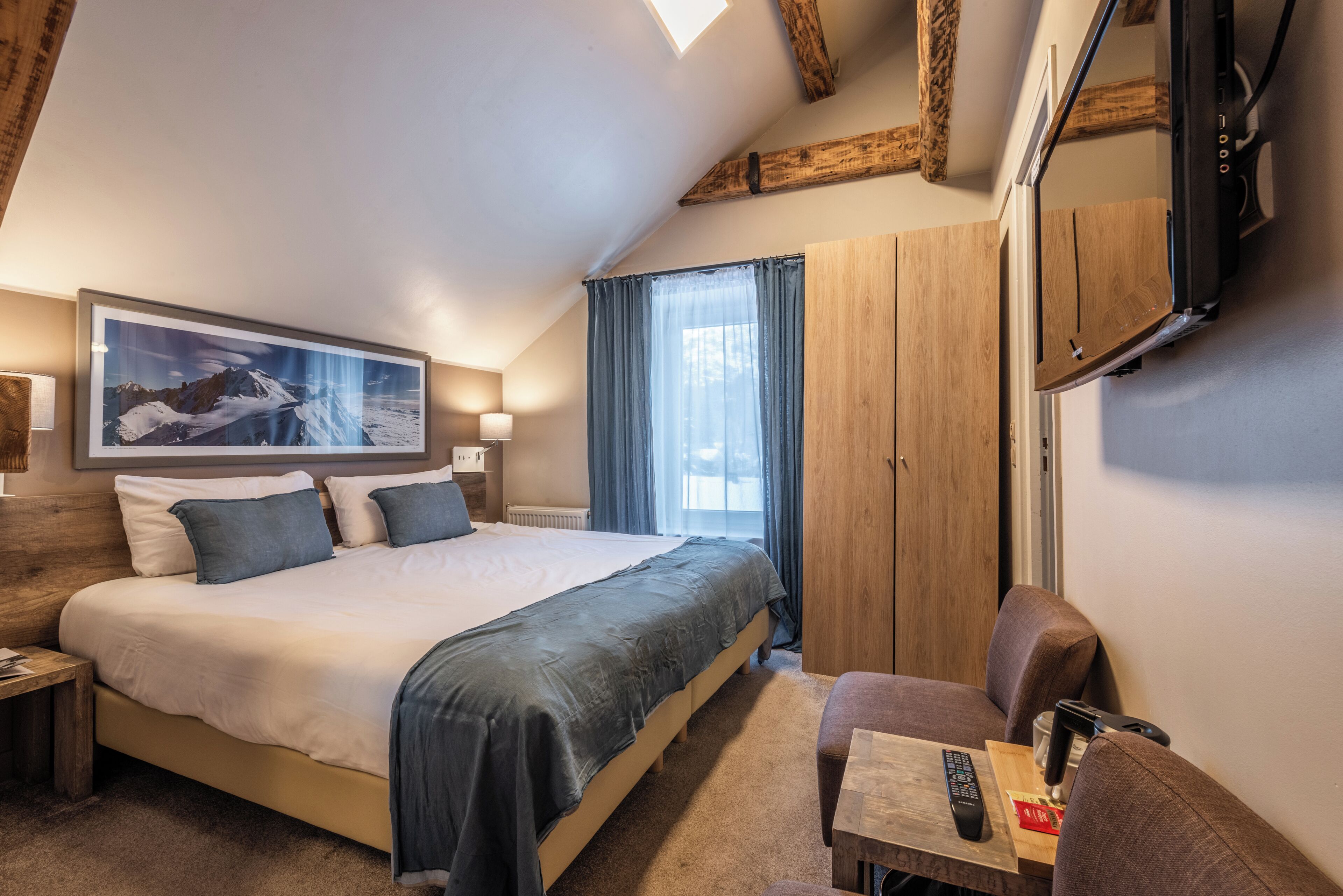 A bedroom with a large bed, a wooden headboard, a mounted TV, a desk with a chair, and a window with curtains.