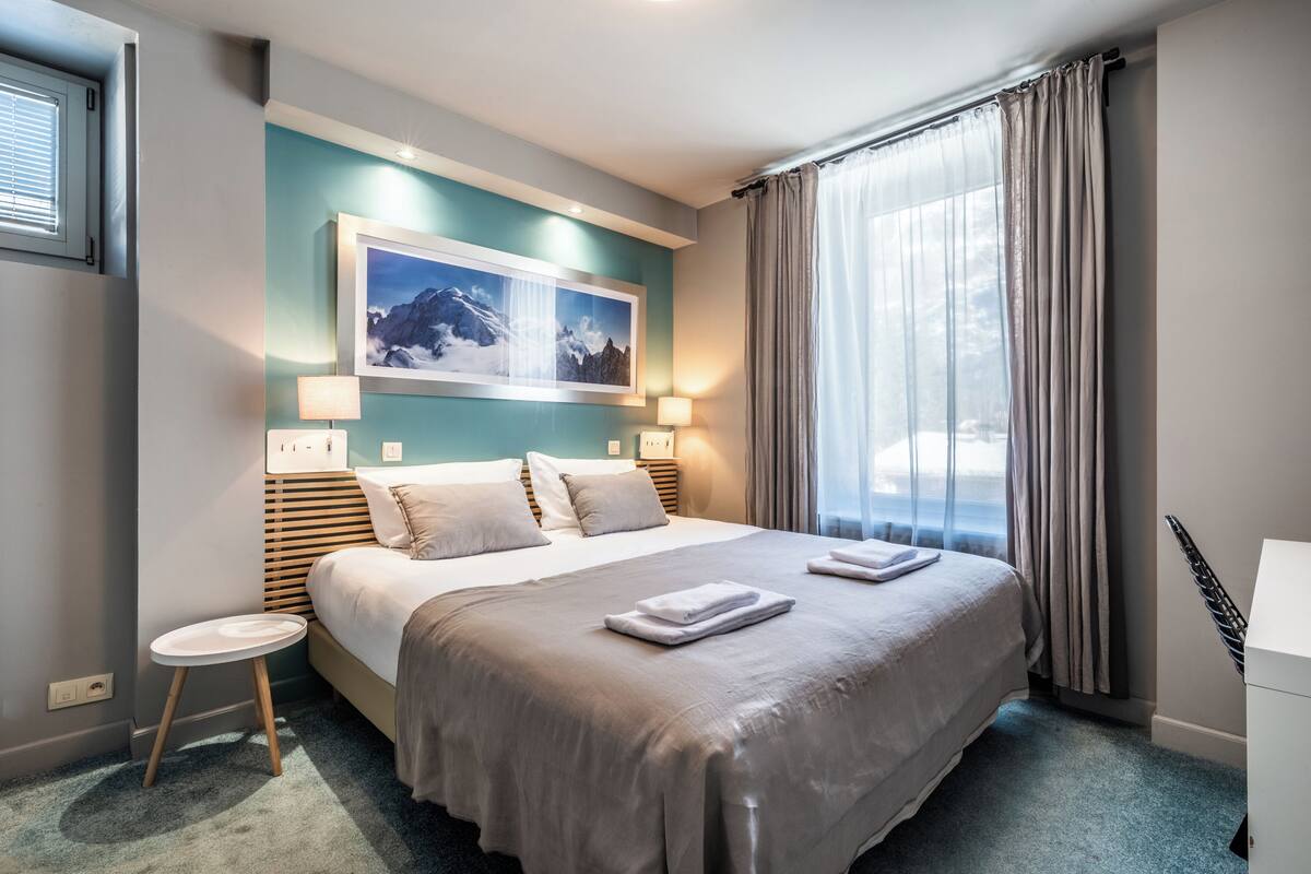 Quarto de hotel moderno com uma cama grande, uma mesinha de cabeceira, um quadro pendurado e vista para o exterior através de cortinas translúcidas.