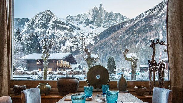 A dining table with chairs and a view of snow-covered mountains through a window. - Hotel Eden Chamonix (Chamonix-Mont-Blanc)