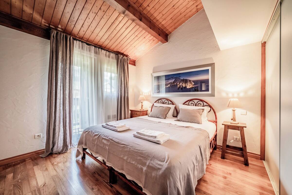 Quarto com teto de madeira, uma cama grande com cabeceira, duas mesas de cabeceira com abajures e um quadro emoldurado na parede.