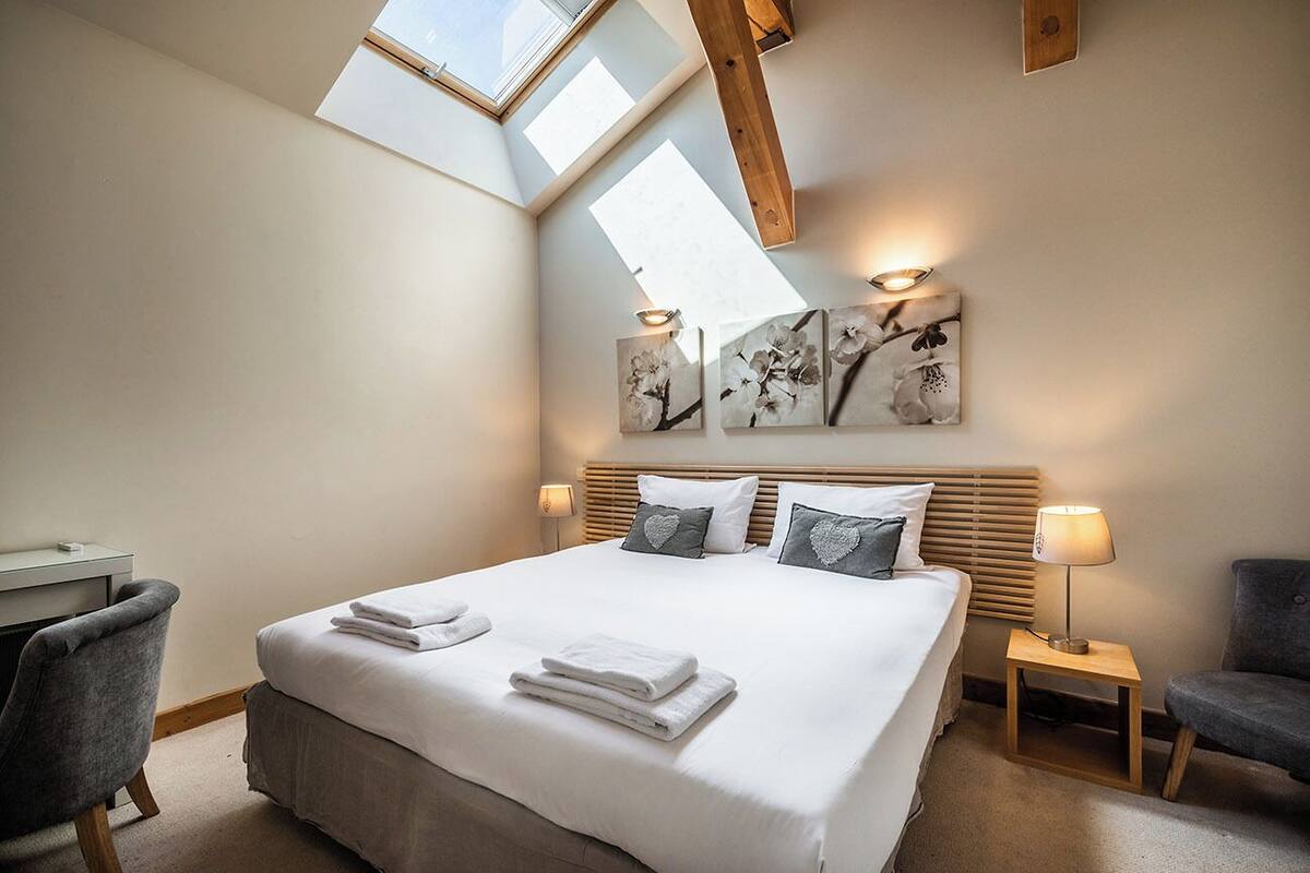 Quarto com cama grande, duas mesas de cabeceira, escrivaninha e cadeira. Há claraboias acima da cama e obras de arte na parede.