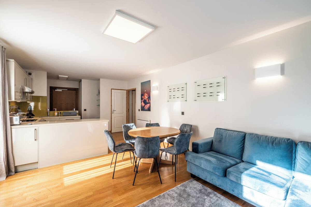 Uma sala de estar moderna com um sofá azul, uma mesa de jantar redonda com cadeiras e uma cozinha ao fundo.