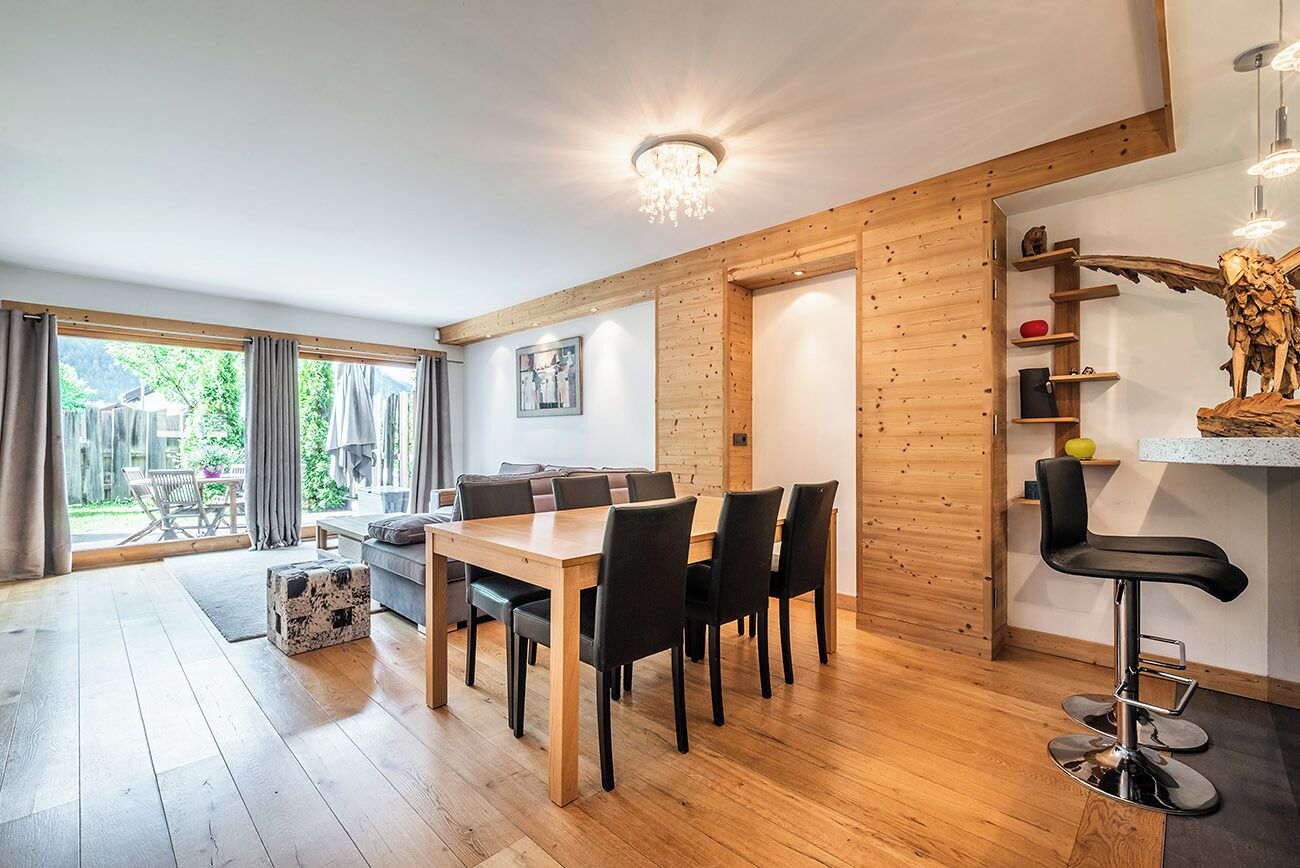 A modern dining area with a wooden table and chairs, a bar stool, and a chandelier.