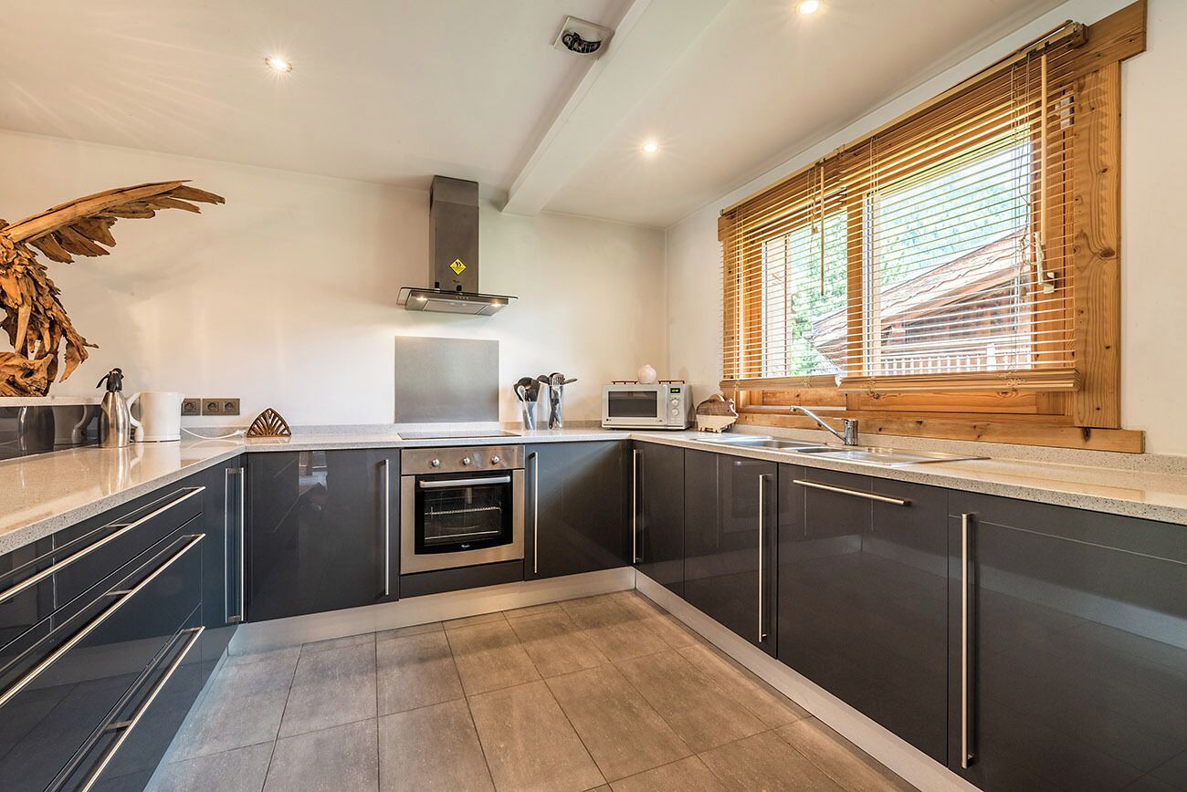 A modern kitchen with dark cabinetry, a built-in oven, a microwave, and a wooden window with blinds.