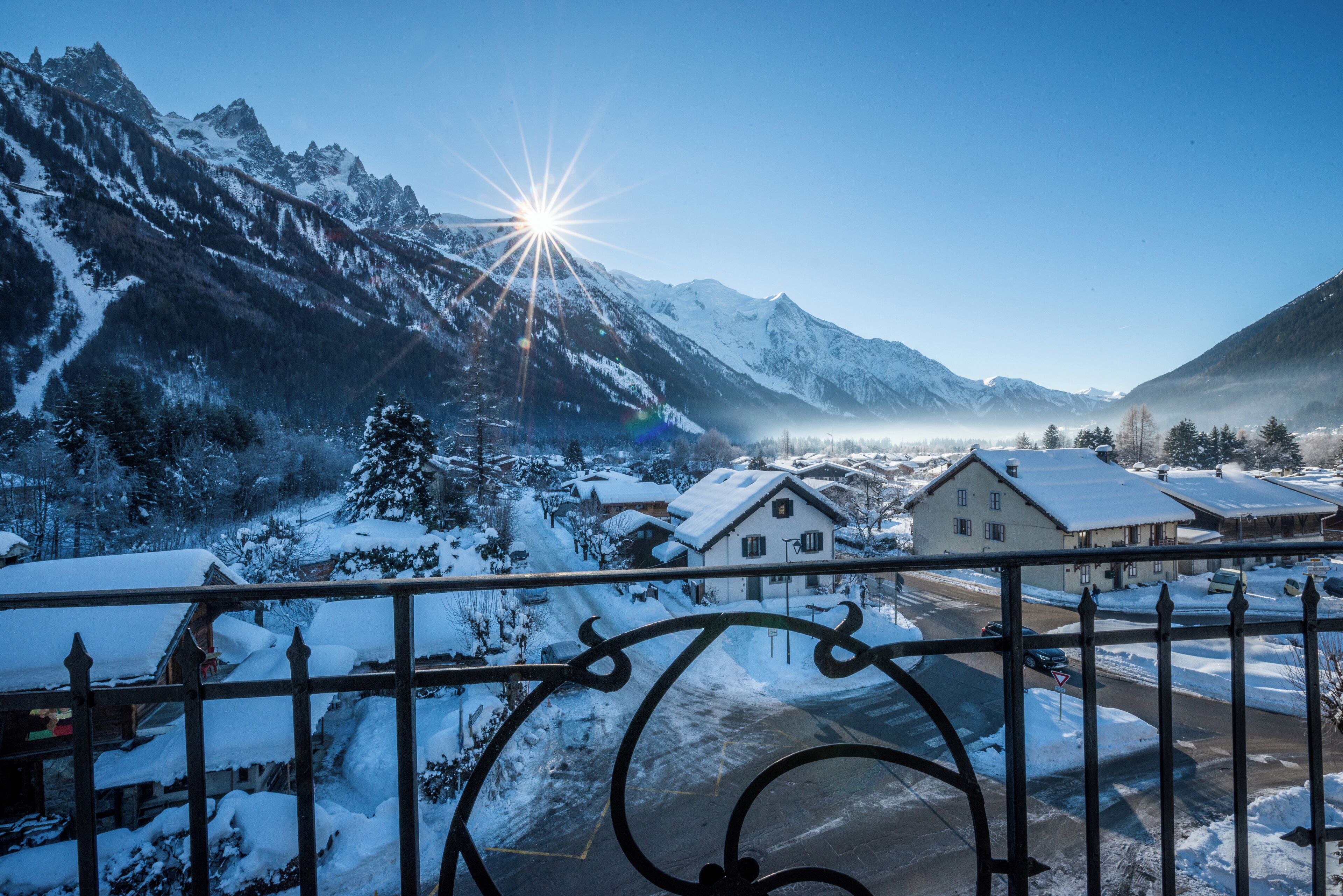 A snow-covered mountain landscape with a clear sky, a sun shining through the peaks, and a residential area with houses and a road in the foreground.