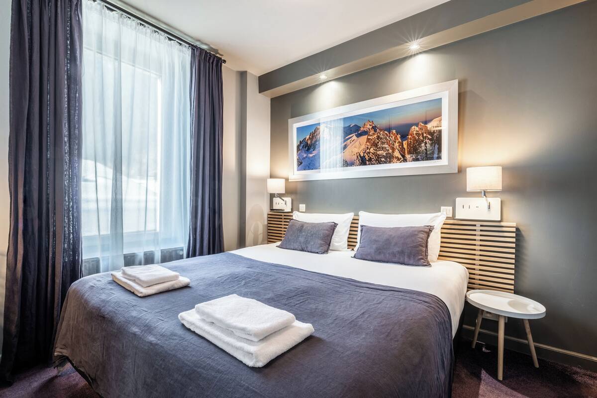 Quarto de hotel com uma cama grande, uma mesinha de cabeceira, um quadro pendurado na parede e uma janela com cortinas.