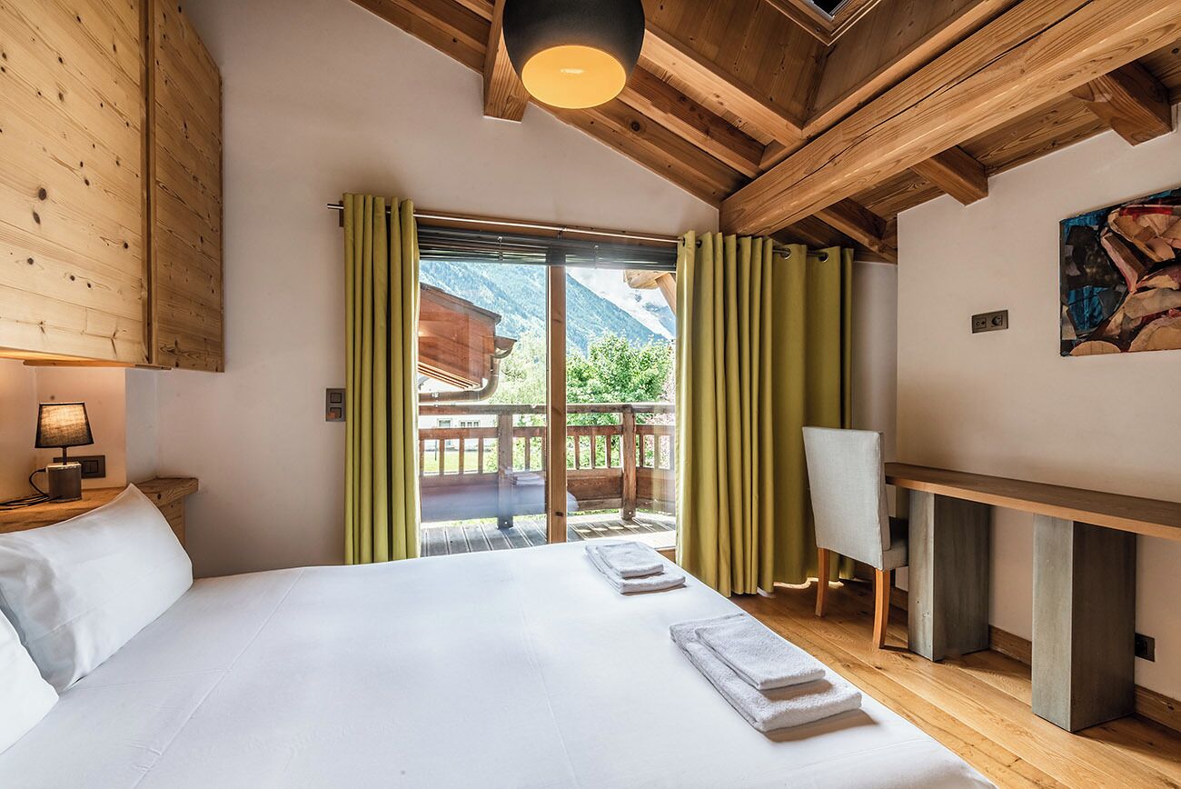 A bedroom with a wooden ceiling, a bed with white linens, a desk, and a view of mountains through a sliding door.