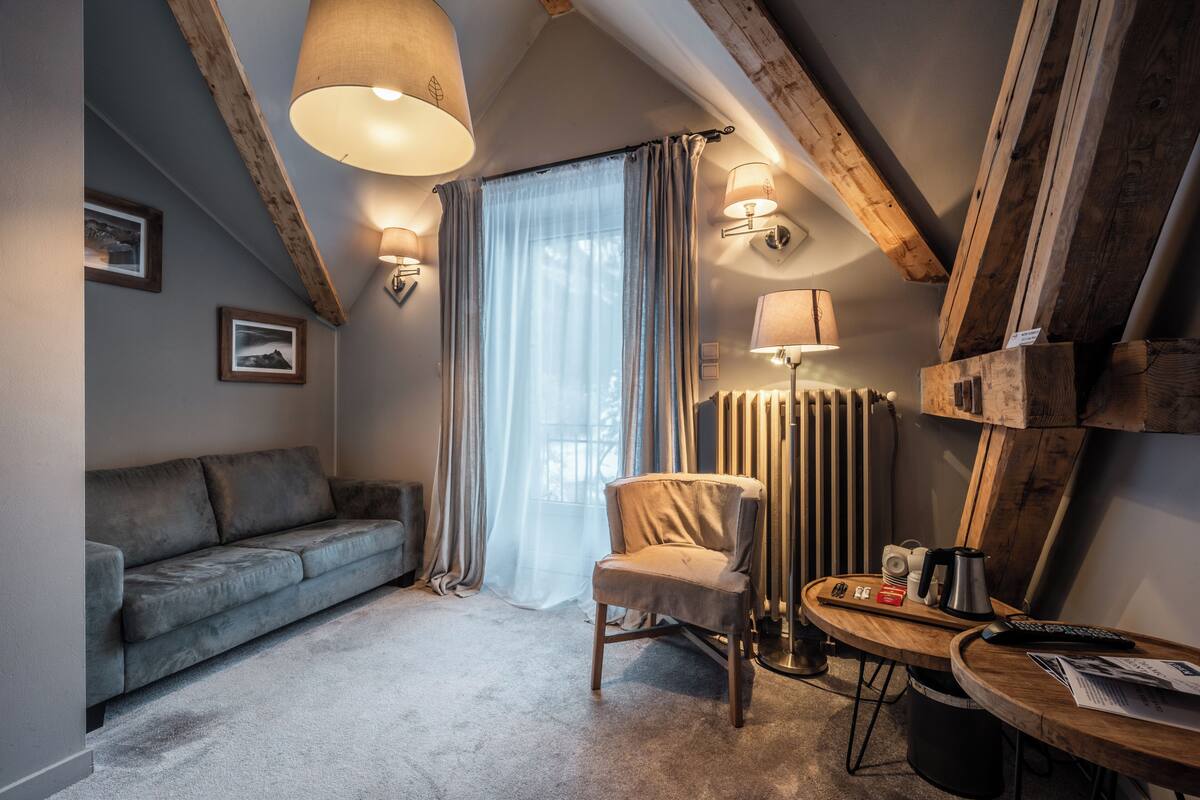 Quarto acolhedor com sofá cinzento, uma cadeira de madeira, uma pequena mesa de madeira com uma chaleira, chávenas e um comando. Existem duas luminárias de parede e um radiador.