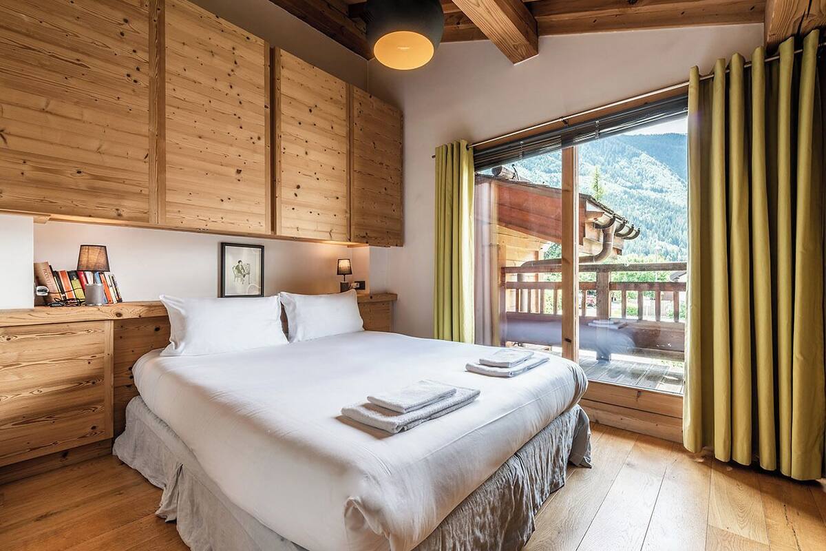 Um quarto com teto de madeira, uma cama grande com roupa de cama branca, uma mesa de cabeceira com abajur e vista para uma montanha pela janela.