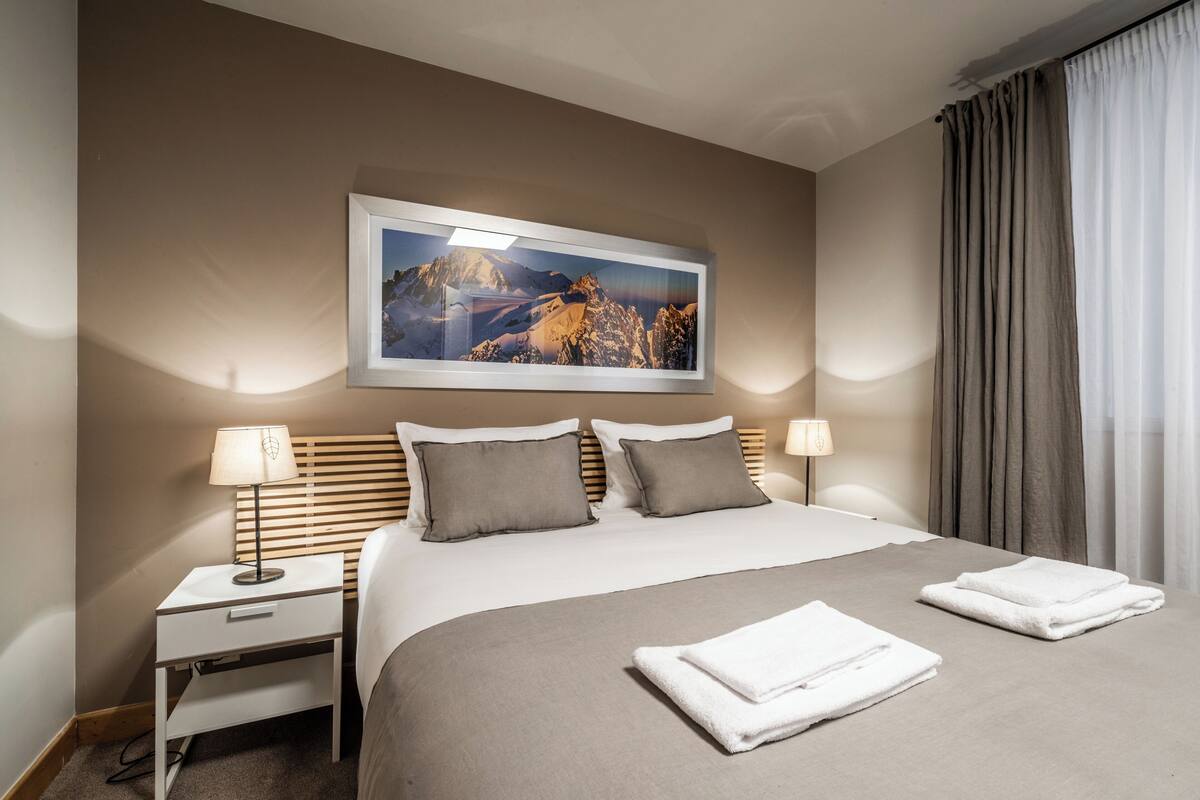 Quarto de hotel com cama, duas luminárias de cabeceira, mesa de cabeceira, quadro de paisagem na parede e janela com cortinas.