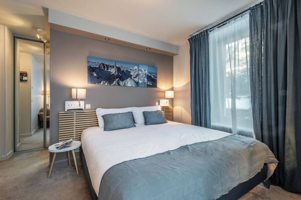 Quarto de hotel moderno com uma cama grande, duas luminárias laterais, uma mesinha de centro com uma revista, um quadro de montanhas na parede e uma janela ampla com cortinas.