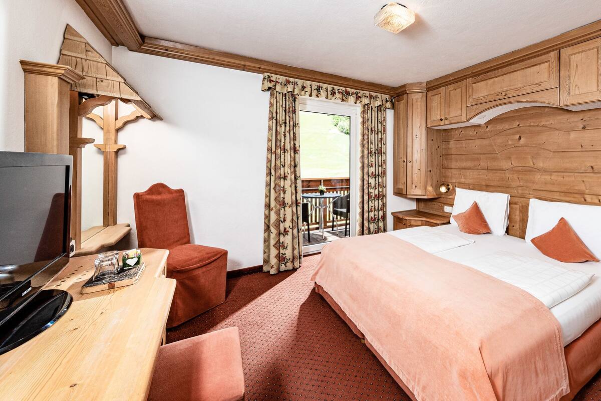 A hotel room with a wooden headboard, a bed with white and orange bedding, a desk with a television, and a view of a balcony with curtains.