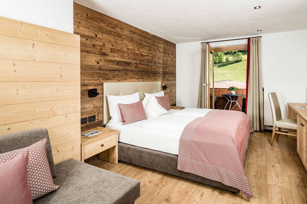 A modern hotel room with a wooden wall, a large bed with patterned pillows, a desk with a chair, and a view of a green landscape through a window.