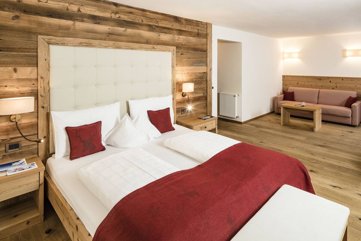A hotel room with a large bed, wooden headboard, bedside tables with lamps, a wooden dresser, a small table, and a sofa.