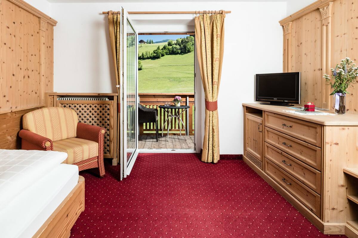 A hotel room with a bed, a chair, a TV, and a view of a green landscape through an open door.