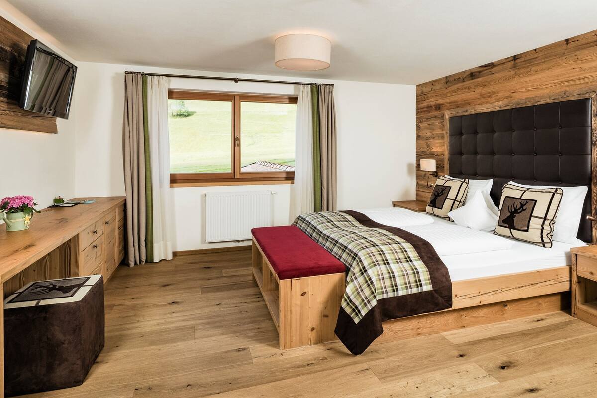 A bedroom with a wooden headboard, a bed with a plaid bedspread, a bench, a desk with a plant, and a window with curtains.
