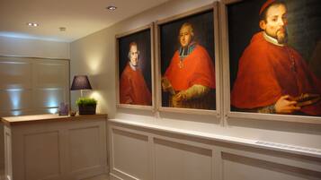 Un couloir orné de trois portraits encadrés représentant des personnes vêtues de robes rouges, ainsi qu’une lampe et une petite plante posée sur une étagère.